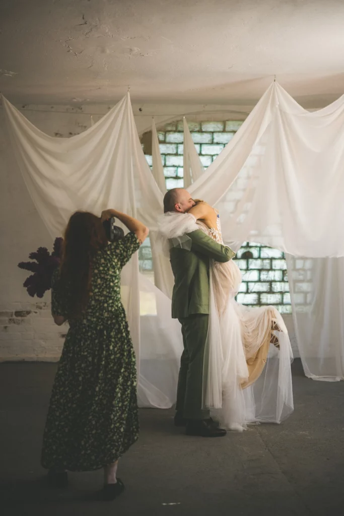 Portfolio fotograficzne Kamila Bryckiego - backstage sesji ślubnej robionej przez duet Nina & Darek. Pan Młody podnosi Pannę Młodą, a fotografka robi zdjęcia.
